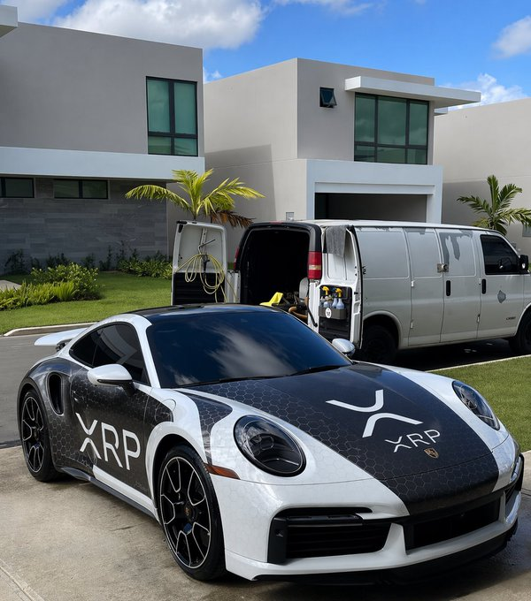 A white Porsche sports car with black hood and "XRP" decals parked in front of a modern house, with a white van and another vehicle visible behind it.