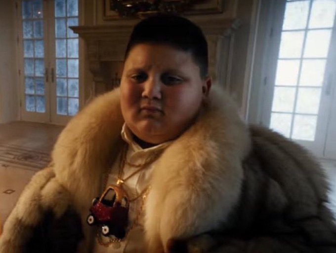 A young boy stands indoors wearing an oversized fur coat, with a small red toy car hanging from a chain around his neck. The lavish image contrasts with the post’s warning that a temporary AI boom would direct immense computing power and investment into profit—prompting viewers to consider who or what benefits.