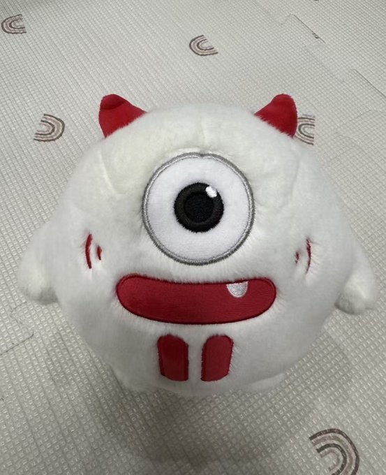 A round white plush toy with red horns, one large eye, a wide red smile with a fang, and small red legs, sitting on a textured white mat.