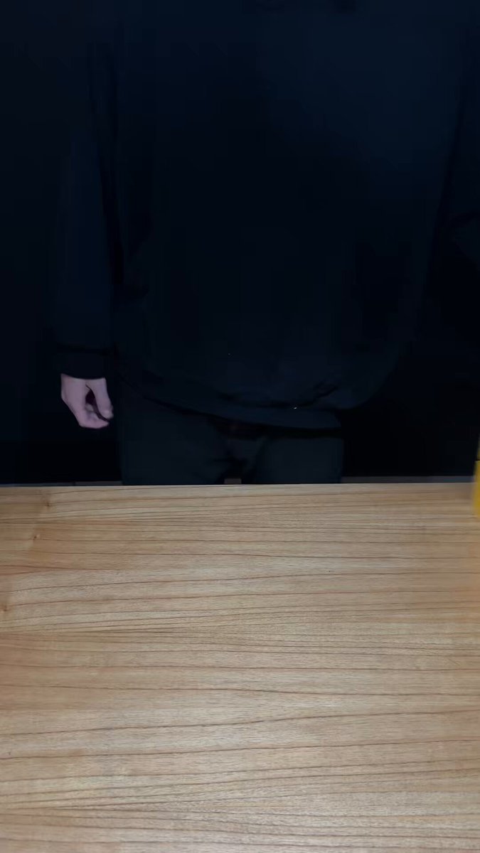 The image features a person standing next to a wooden table with a brown surface. The person has their back turned towards the camera, making them appear as if they are facing away from us. They are wearing black clothing, which contrasts with the light wood grain of the table. In the background, there is a yellow object that stands out against the dark backdrop.