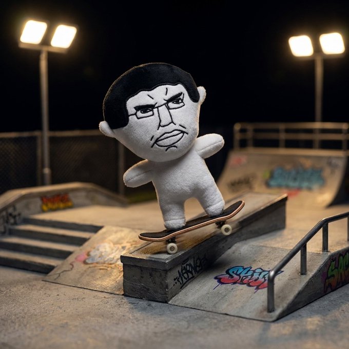 A plush figure with a stern-looking face riding a miniature skateboard on a ramp at a tiny skatepark setup at night.