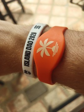 The image shows two wristbands, one orange and one white, worn by a person's arm. The orange band has a palm tree design on it, while the white band features a black and white logo. Both bands are positioned close together on the wrist of the person wearing them.