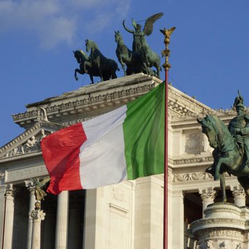 An Italian flag (green, white, and red) waving in the foreground against a backdrop of classical architecture featuring bronze equestrian statues and winged figures under a blue sky.