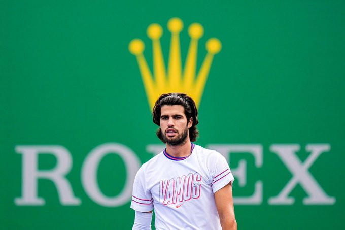 A bearded tennis player in a white shirt stands in front of a green backdrop featuring the Rolex crown logo and 'ROLEX' text.