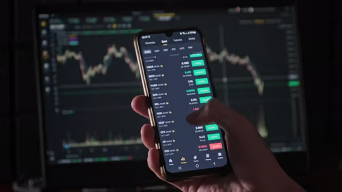 A hand holds a smartphone displaying a crypto trading app with green and red price movements and order book data, set against a desktop monitor showing candlestick charts. The image reinforces the post's message about obsessively checking charts during the holiday—capturing the lone trader glued to market data while the rest of the world enjoys the 4th of July.
