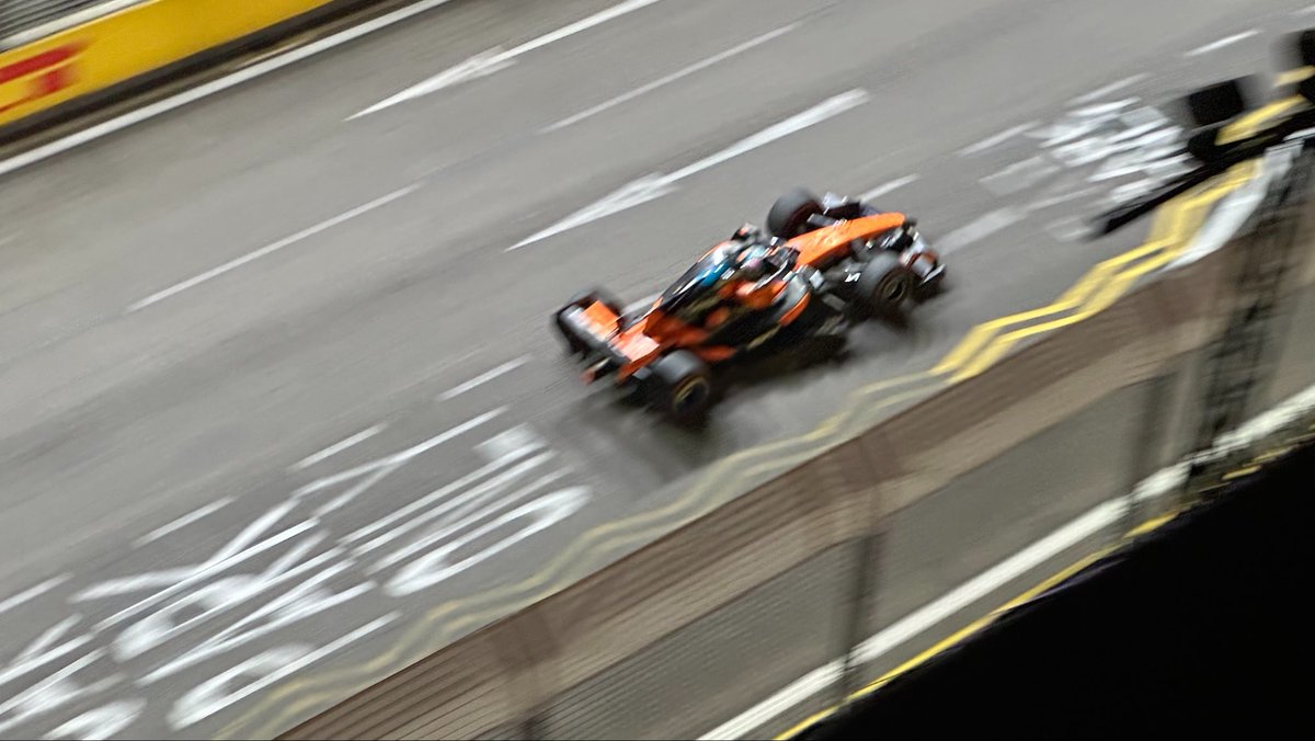 A Formula 1 car speeds along a track, captured with motion blur that emphasizes its velocity, viewed from an elevated vantage point overlooking one of the circuit's long straits. The dynamic shot illustrates the high-speed racing experience the post describes from the OKX F1 viewing area atop the National Gallery, with the iconic Marina Bay Sands likely providing the elevated perspective.