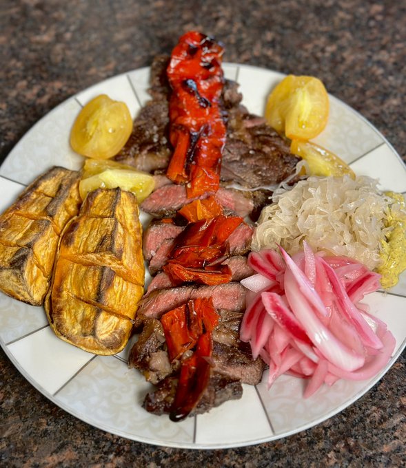 A white patterned plate containing sliced steak topped with grilled red bell peppers, accompanied by grilled eggplant slices, yellow pickled peppers, sauerkraut, and sliced pickled red onions.