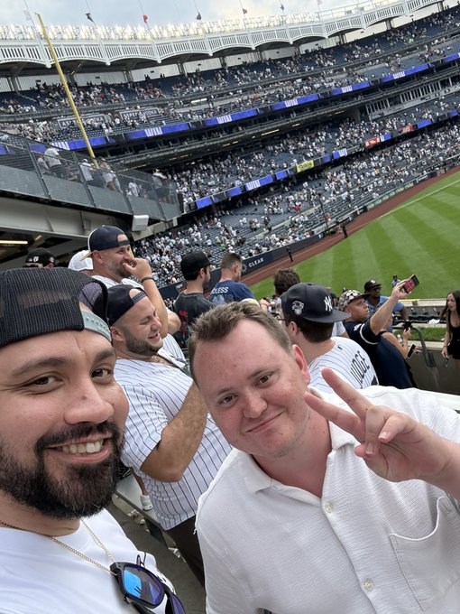 Two men take a selfie while attending a New York Yankees baseball game at Yankee Stadium, with the pinstripe jerseys and 'NEW YORK' signage visible in the stands behind them.