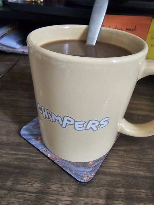 A beige ceramic mug labeled "CHIMPERS" filled with coffee and a spoon, sitting on a coaster on a wooden table.