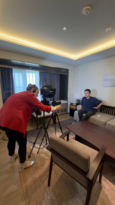 A woman in a red jacket operates a camera on a tripod to film a man sitting on a beige sofa in a hotel room or apartment.
