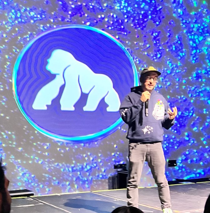 A man wearing a blue hoodie and cap stands on a stage with a microphone, presenting in front of a large screen displaying a glowing blue and white gorilla logo.