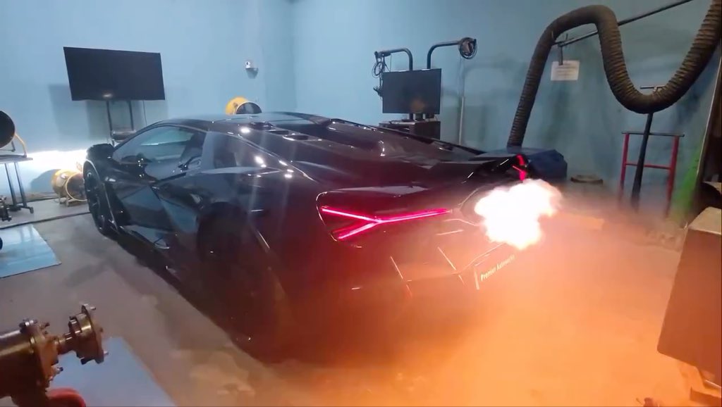 Black sports car (appears to be a Lamborghini Revuelto) on a dynamometer in a garage, with flames shooting out of the exhaust and smoke filling the room.