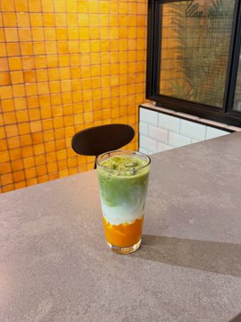A tall glass containing a layered green and orange beverage sits on a gray table against a vibrant yellow tiled wall background.