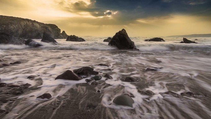 A moody coastal landscape photograph showing waves washing over a rocky beach at sunset or sunrise with golden light breaking through clouds.
