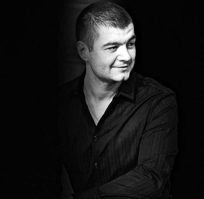 A man with short hair, wearing a dark button-up shirt and a necklace, smiles subtly while looking off to the side in a black-and-white portrait. The accompanying post suggests he is sharing a personal health update about a recent leukemia diagnosis, lending a poignant, reflective tone to the image.
