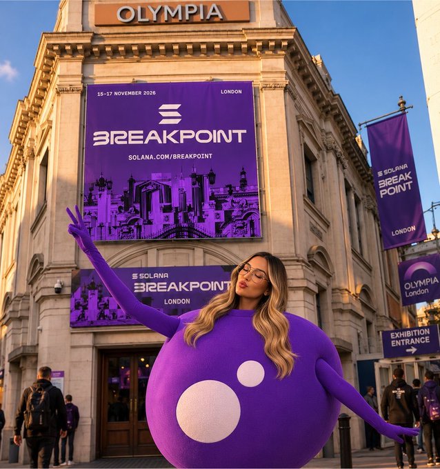 A woman in a purple inflatable costume with a peace sign poses in front of London's Olympia venue, which is wrapped in "Solana Breakpoint" banners advertising the event on 15-17 November 2026 (solana.com/breakpoint). The image ties to the caption by showing the excited announcement that the creator is officially attending the Solana Breakpoint conference in London, sponsored by MoonPay.