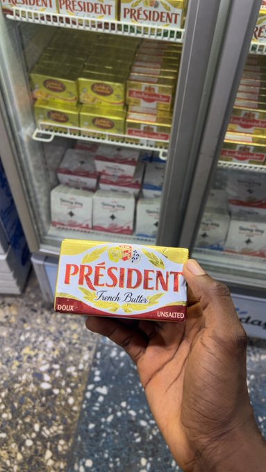 A hand holding a package of Président French Butter (unsalted) in front of a refrigerated display case stocked with various butter products.