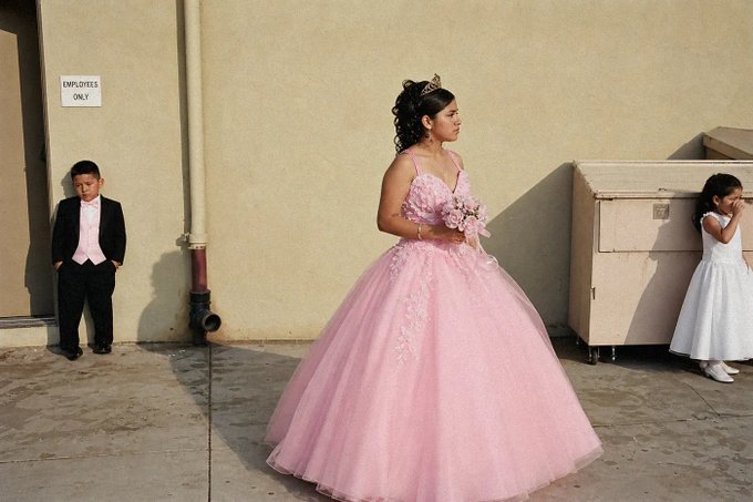 A young woman in a pink princess gown and tiara stands between a boy in a black tuxedo and a girl in a white dress, posing like royalty during what appears to be a quinceañera. An "EMPLOYEES ONLY" sign is visible on the wall behind them.