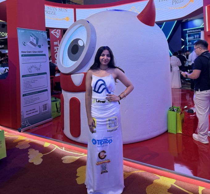 A young woman wearing a strapless dress covered in tech company logos poses in front of a large mascot character at an event booth, with a sign partially reading 'How Can I Use 1NCE?' visible to the left.