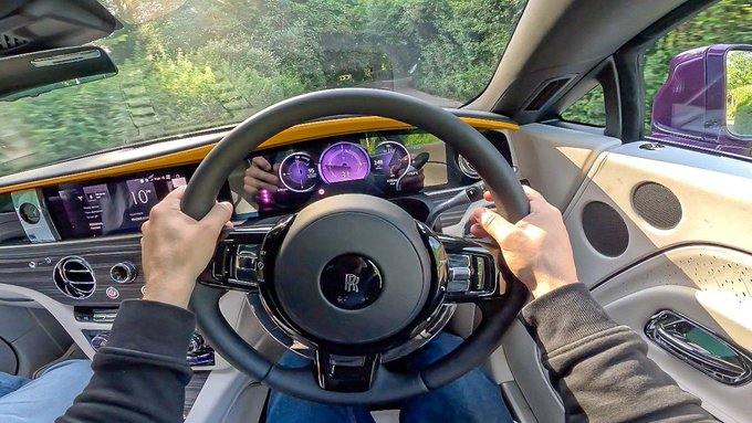 First-person view from the driver's seat of a Rolls-Royce, showing hands on the steering wheel with a scenic road and greenery visible through the windshield.