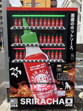 A Japanese Coca-Cola Sriracha vending machine stocked with red bottles, labeled "悪魔のホットソース" (Devil's Hot Sauce), with Sriracha branding and flames imagery at the bottom, located on a street.