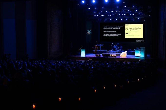 A dimly lit theater stage displays a presentation slide reading "Know the ecosystem before you build" during what appears to be a tech event, with a silhouetted audience facing a speaker illuminated by blue spotlights.

The image aligns with the post's description of a large gathering (approximately 1,500 attendees) at a provincial theater event featuring presentations on the Cursor AI ecosystem, smart contract development with Celo, and Real World Assets tokenization opportunities.