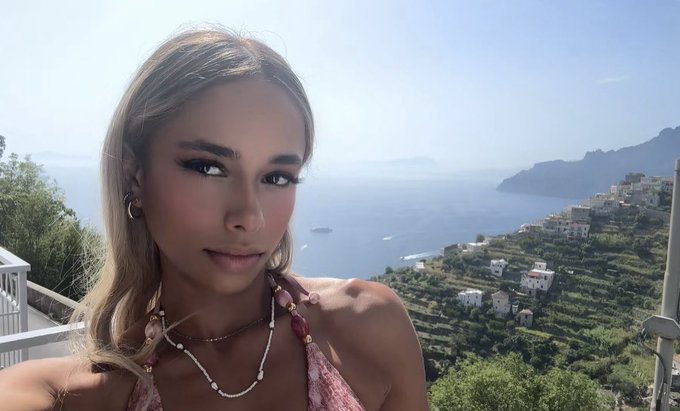 A young woman with blonde hair takes a selfie with a stunning coastal view behind her, showing the sea and hillside buildings of what appears to be the Amalfi Coast.