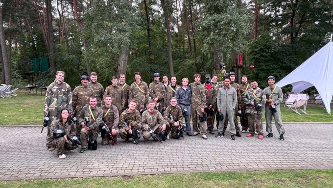 A group photo of approximately 20 people, mostly in military camouflage uniforms, posing outdoors on a brick patio with trees and a white tent in the background.