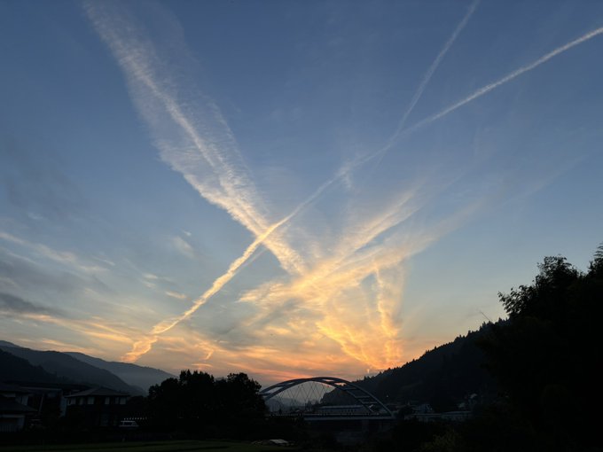 A landscape photograph at dusk showing a dramatic sky filled with crisscrossing jet contrails over a silhouetted valley with mountains and a bridge.