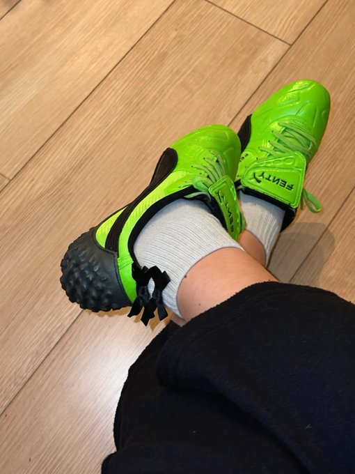 A person lying down wearing bright green Pemtry athletic cleats with matching socks on a wooden floor.