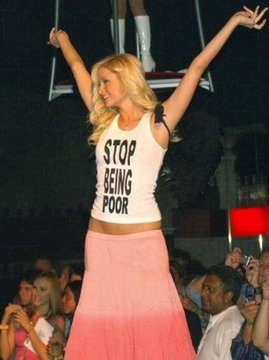 A blonde woman on stage wearing a white tank top with the text 'STOP BEING POOR' paired with a pink ombre skirt celebrates with her arms raised.