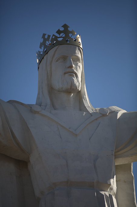 A large white stone statue depicting a bearded figure wearing a crown, photographed from a low angle against a clear blue sky.
