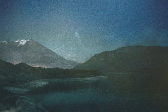 A moody nighttime photograph of a mountain landscape with a body of water, dark silhouettes, and a faint hint of the Milky Way in the starry sky.