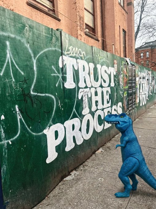 A small blue toy dinosaur stands on a city sidewalk in front of a green construction hoarding bearing the graffiti slogan "TRUST THE PROCESS," visually echoing the post's message about trusting the journey while buying more of the ticker $fefer.