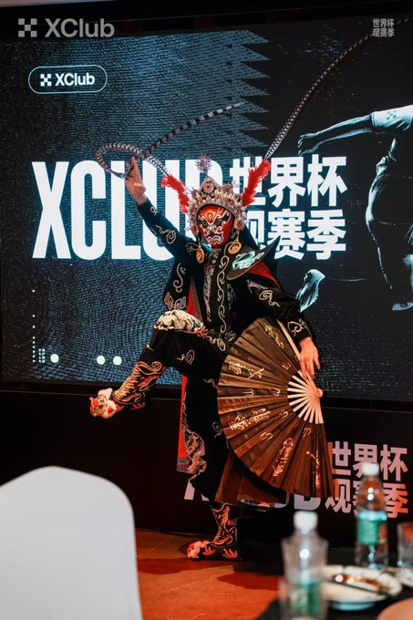 A performer in elaborate traditional costume and ornate headdress executes a dynamic pose with a large folding fan at the XClub World Cup viewing season event, as indicated by the "XClub" logo and "XClub 世界杯 观赛季" text on the screen behind him, showcasing the mesmerizing Sichuan Opera face-changing art.