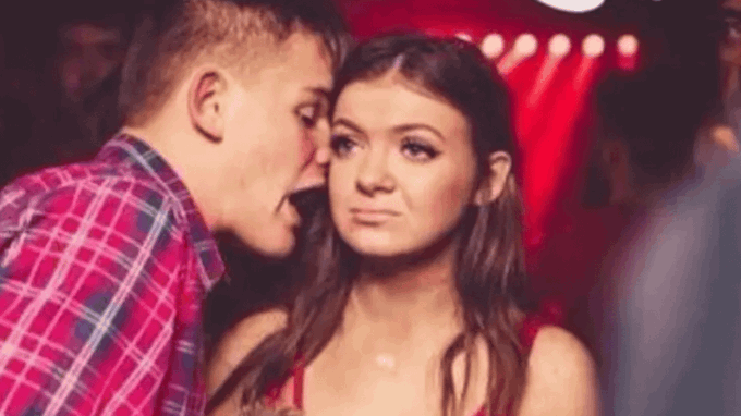 A young man in a red plaid shirt whispers into the ear of a young woman in a red top at what appears to be a club with red lighting in the background.