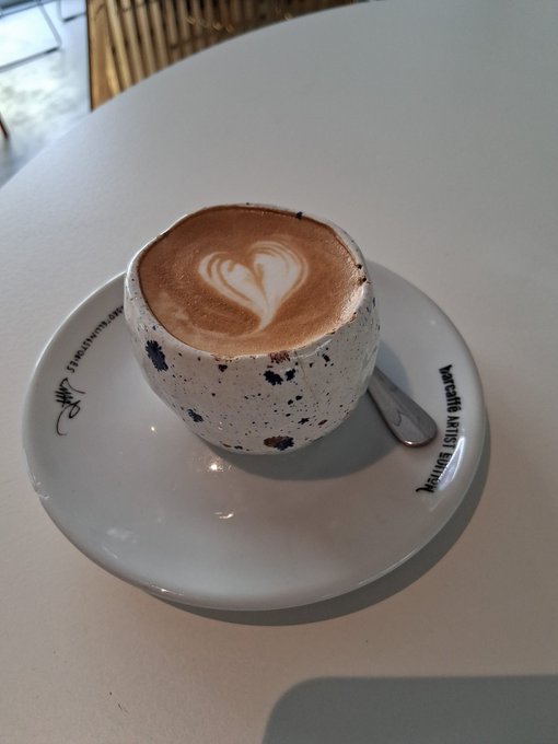 A cappuccino with a heart-shaped latte art served in a speckled ceramic cup on a saucer reading 'Mercantile Arts (IST) Boutique'.