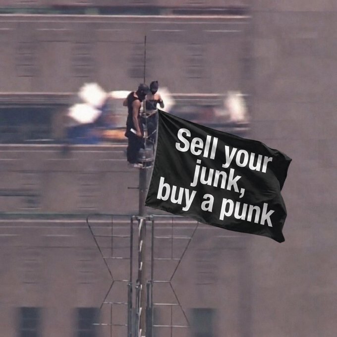 A photograph of two workers on a tall antenna/broadcast tower in a city, with a large black flag overlay reading 'Sell your junk, buy a punk' — a play on the iconic 'Jolly Roger' CryptoPunk NFT flag scene.