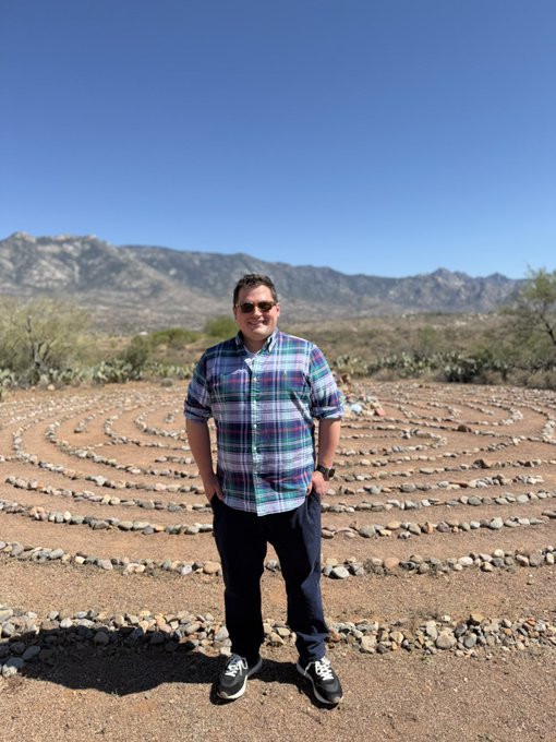 A man in a plaid shirt and sunglasses standing in front of a stone labyrinth in a desert landscape with mountains in the background.