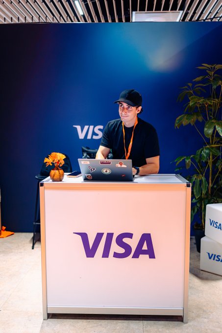 A man in a black cap and shirt sits behind a Visa-branded counter, working on a laptop, with a blue Visa backdrop and a potted plant beside him.