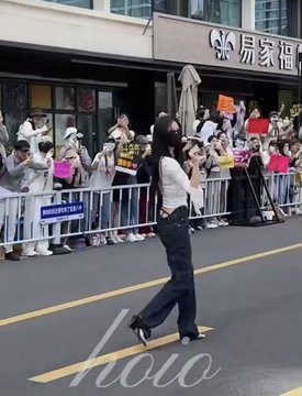 Street photo of a young woman in a white top and dark flared pants posing on a road with arms raised, in front of a barricaded crowd of fans holding signs during what appears to be a celebrity event in China, with 'hoho' watermark visible.