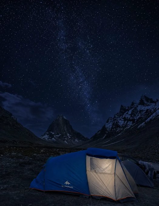 A nighttime landscape photograph featuring an illuminated blue Quechua tent in the foreground, set against a starry sky with the Milky Way and snow-capped mountains.