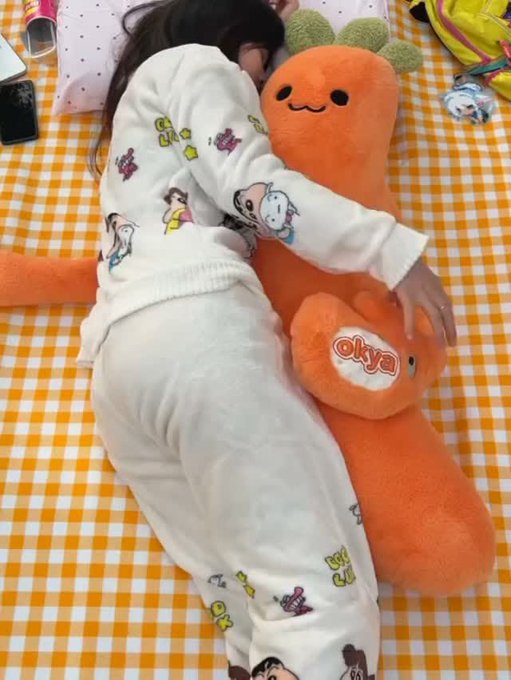 A person in white pajamas with colorful cartoon character prints lies face-down on a yellow and white checkered blanket, hugging a large orange carrot plush toy labeled 'okya'.