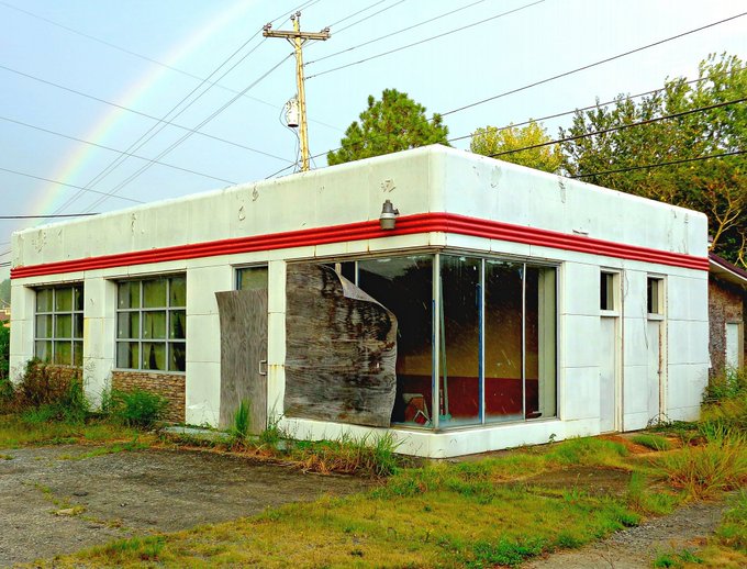 An abandoned vintage white building with red trim, featuring a broken storefront window covered by wood, with a rainbow visible in the sky behind it.