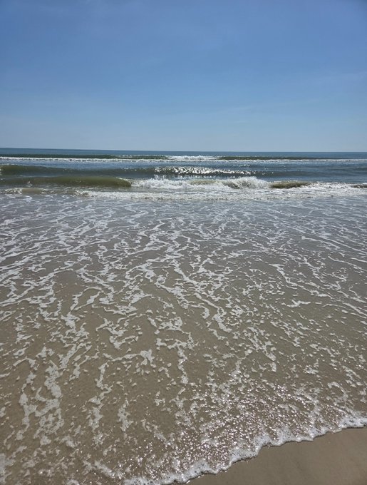 A bright, sunny view of gentle ocean waves rolling onto a sandy beach under a clear blue sky.