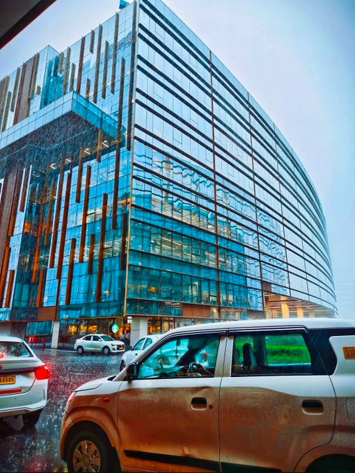 A modern glass building with a blue-green reflective facade, with cars parked in front and rain visible on the foreground vehicle.