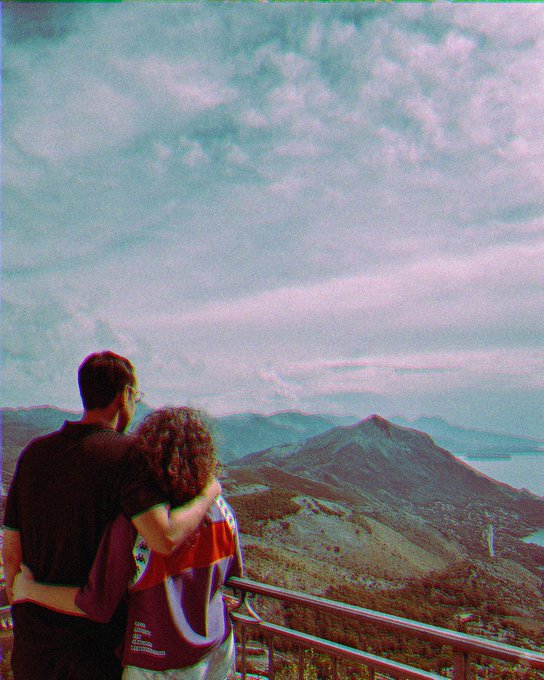 A couple stands on a balcony overlooking a vast landscape of mountains and a body of water under a cloudy sky, with a pink and cyan vintage filter effect.