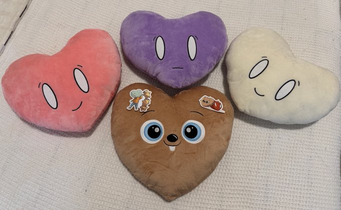 Four heart-shaped plush pillows in pink, purple, brown, and cream colors with simple cartoon faces, including a brown one decorated with stickers, arranged on a white textured surface.
