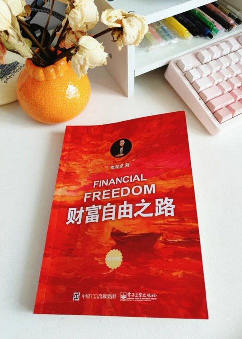 A red-covered book titled "财富自由之路" (Financial Freedom) by 李笑来 (Li Xiaolai) rests on a white desk beside a keyboard and an orange vase with dried flowers, visually accompanying a post reflecting on re-reading Li's earlier work on financial freedom as a personal milestone rather than an endpoint.