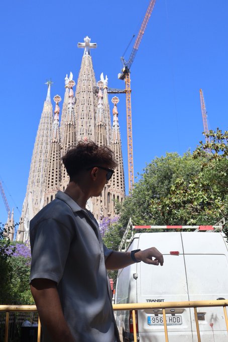 A young man in sunglasses stands in front of the Sagrada Família in Barcelona, with a white Ford Transit van (license plate 9566 HDD) behind him, capturing a sunny travel moment to greet the morning. The iconic basilica and bright blue sky perfectly match the upbeat "GM" energy of the post.